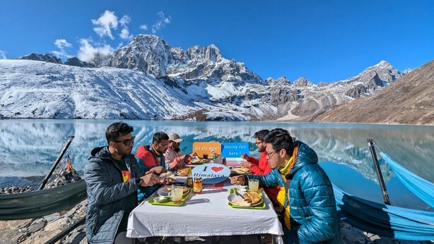 Un grupo de personas cenando en una mesa junto a un lago con montañas al fondo.