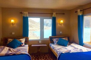 Quarto com duas camas, cada uma com colcha e almofadas azuis, uma mesinha e duas luminárias de parede.
