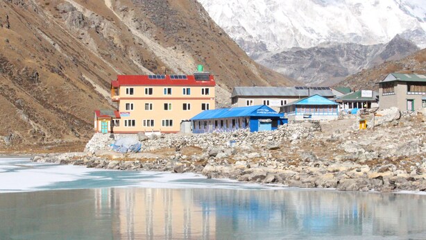 A building with multiple floors and a red roof, situated near a rocky shore with a body of water.