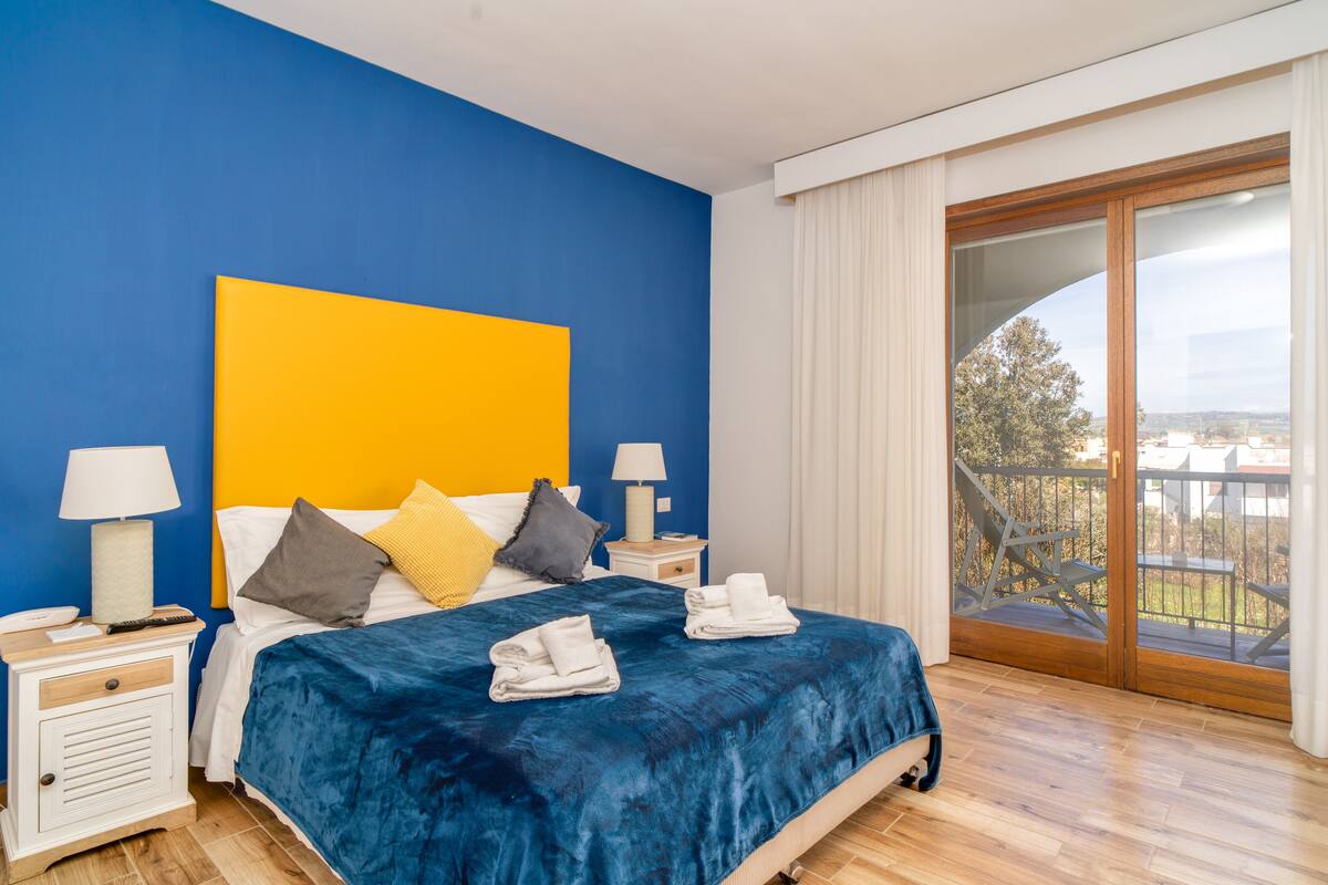 A bedroom with a blue wall, a yellow accent wall, a large bed with a blue cover, two bedside tables with lamps, and a view of the outdoors through a sliding glass door.