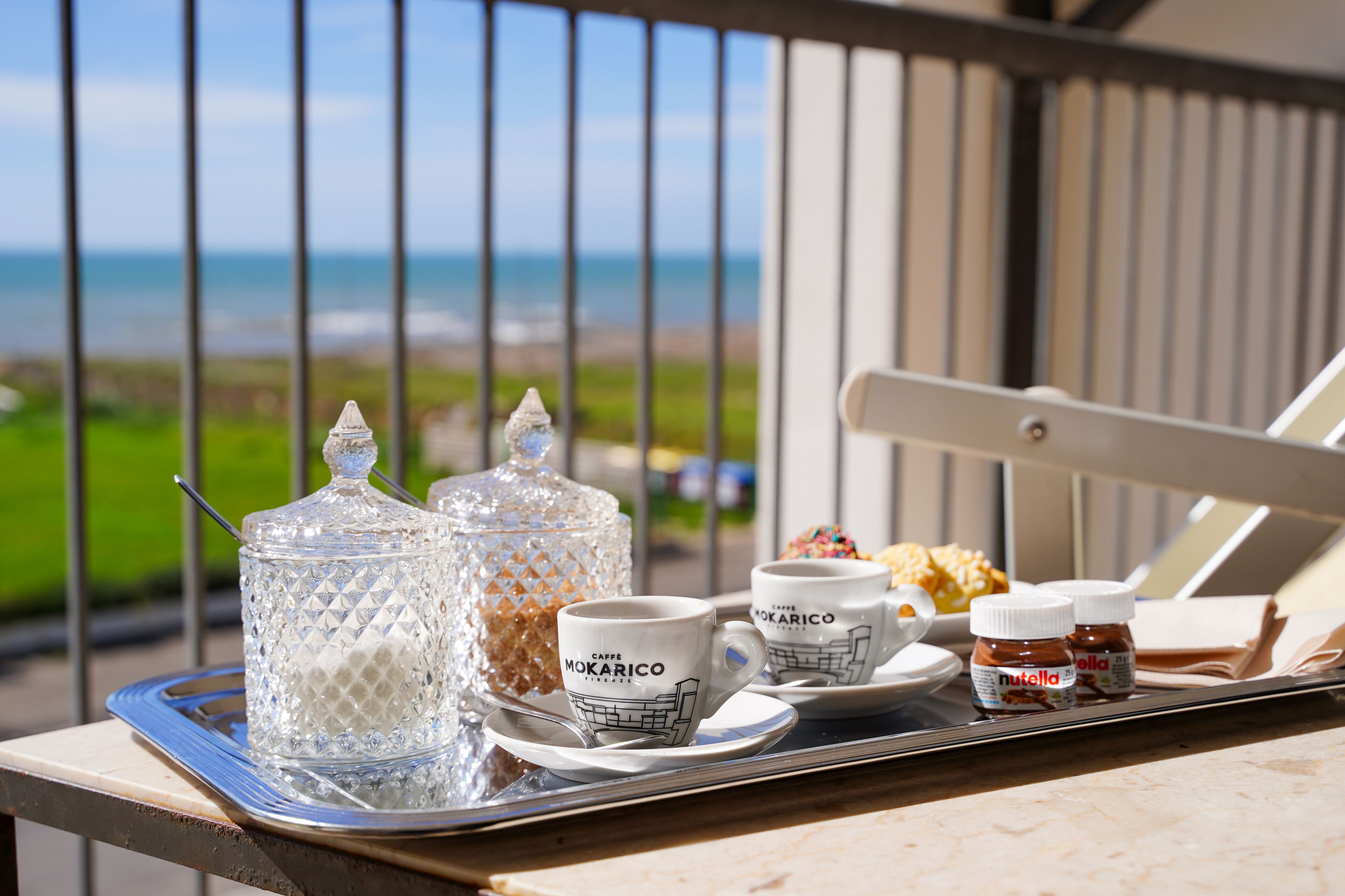A tray with coffee cups, a sugar container, and jars of Nutella on a balcony overlooking a beach.