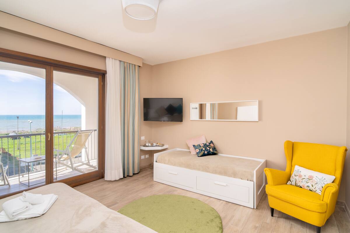 A hotel room with a bed, a yellow armchair, a TV, and a balcony with a view of the sea.