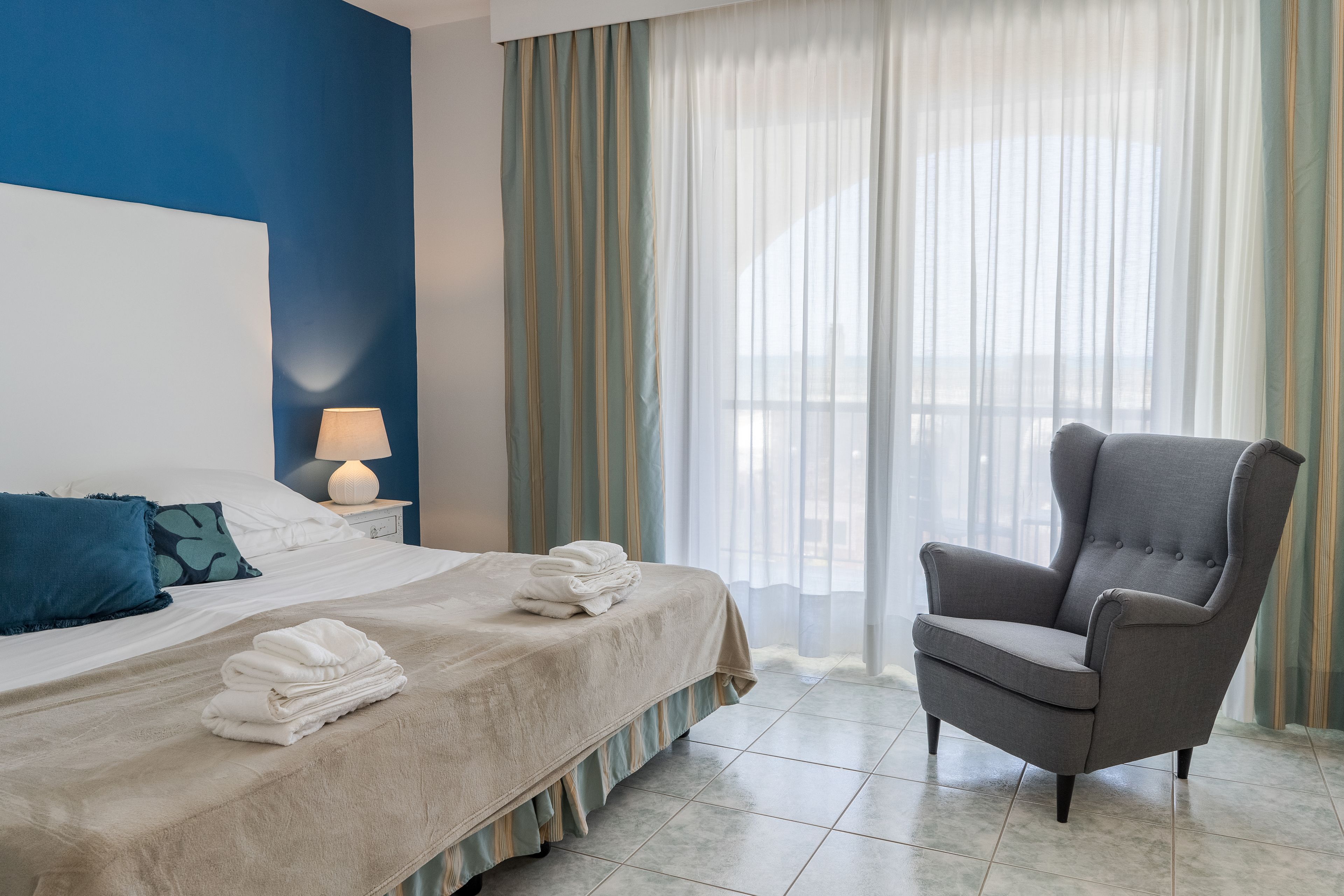 Ein Hotelzimmer mit einem Bett, einem Sessel, einem Nachttisch mit Lampe und einem Fenster mit Vorhängen.