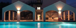 Zona de piscina moderna con vista despejada, área de descanso cubierta y entrada a un edificio.