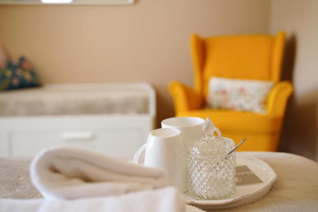 Una habitación ordenada con una cama blanca, un sillón amarillo y una bandeja con un recipiente de vidrio y dos tazas blancas.