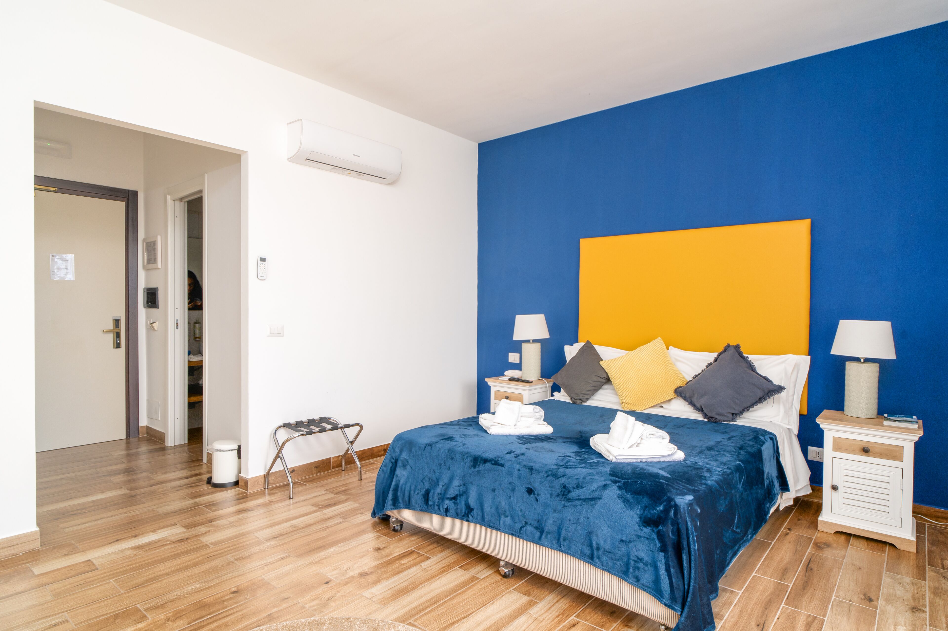 Un dormitorio con una pared azul decorativa, suelo de madera, cama con cubrecama azul, dos mesitas de noche con lámparas y un aparador blanco.