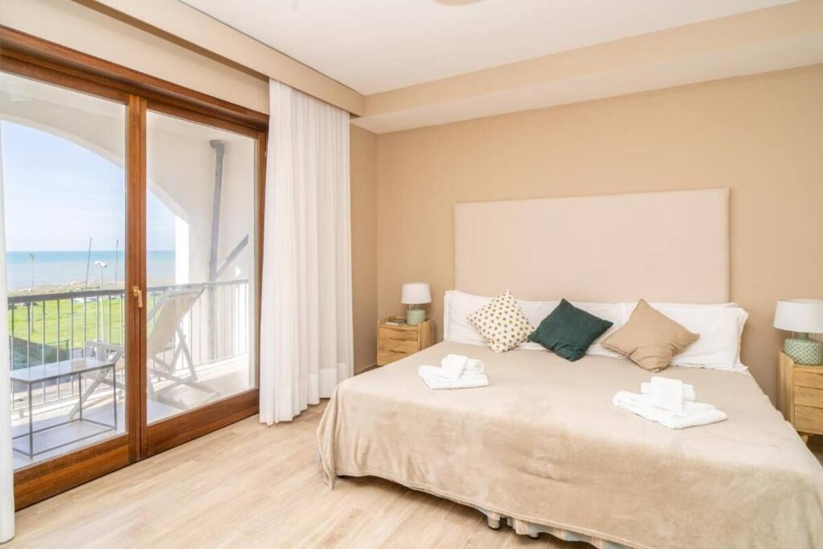 A hotel room with a large bed, two bedside tables, a view of the sea through a sliding glass door, and a balcony with outdoor furniture.