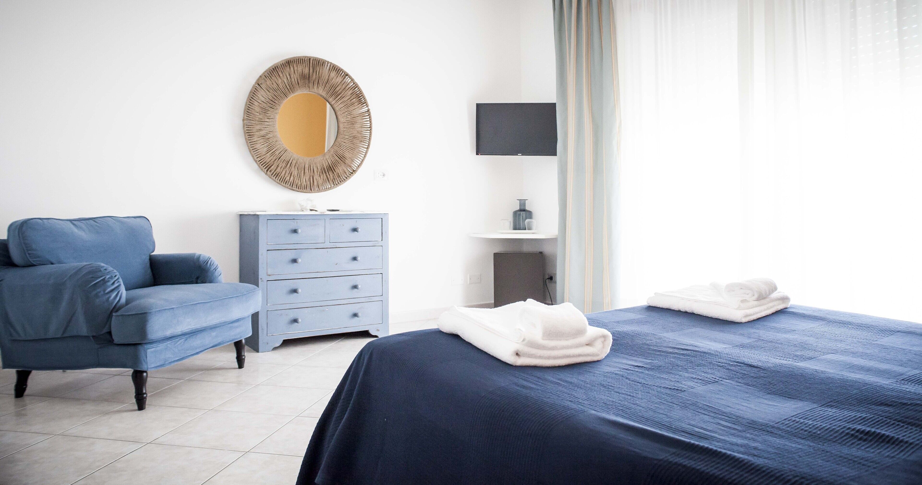 A bedroom with a bed, a blue sofa, a dresser, a mirror, a TV, and a window with curtains.