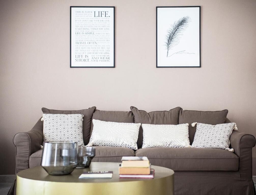A living room with a brown sofa, patterned pillows, a round coffee table with books and a glass, and two framed inspirational quotes on the wall.