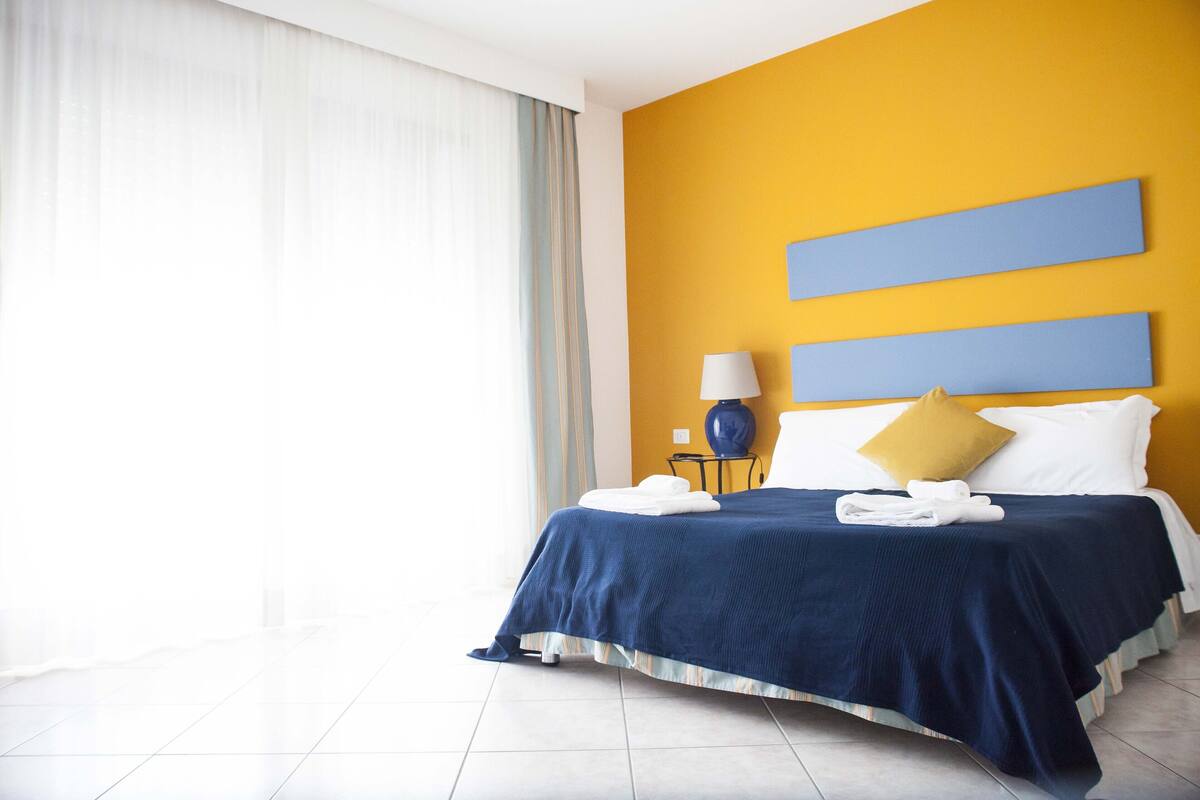 A hotel room with a bed, a nightstand, a lamp, and a window with curtains.