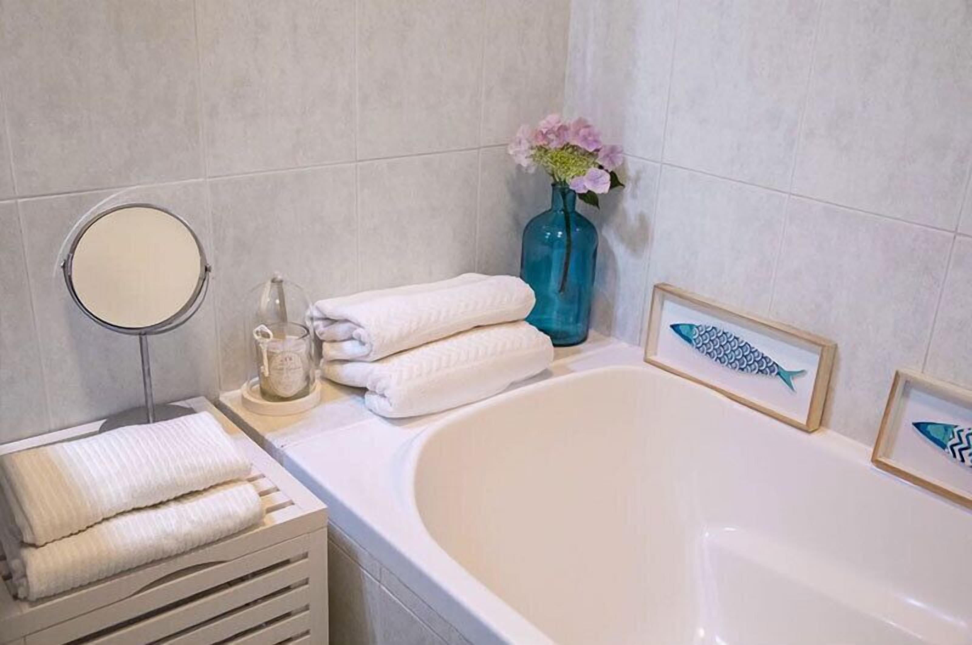 Un baño con bañera, un espejo redondo, un florero con flores y cuadros con motivos de peces.