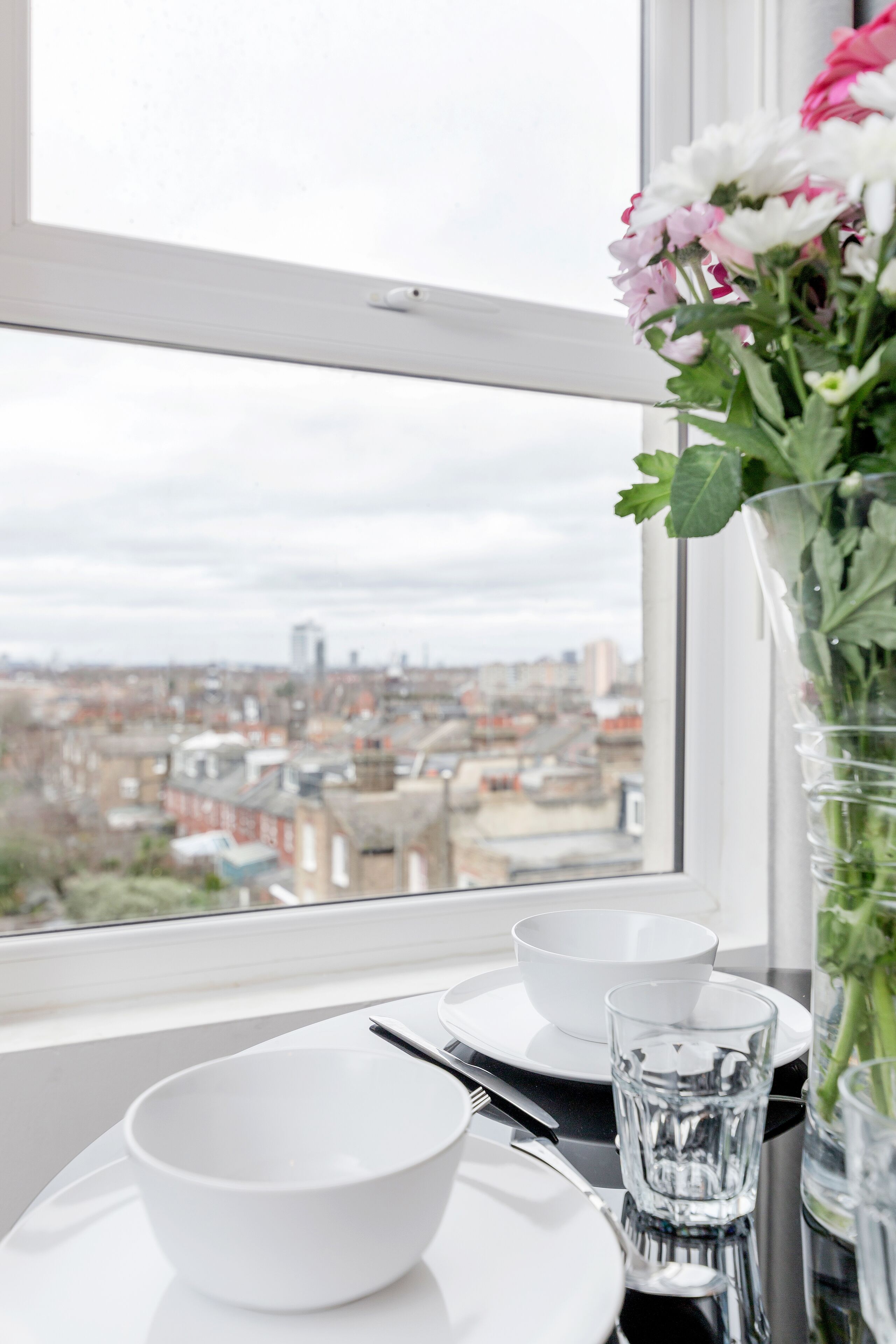 Une table à manger avec des bols blancs, un verre et un couteau, le tout placé devant une fenêtre offrant une vue sur la ville.