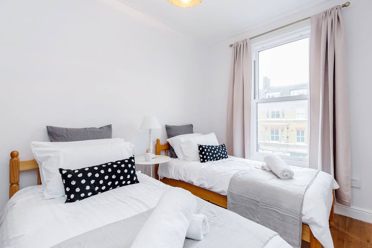 Een hotelkamer met twee enkele bedden, een houten hoofdbord, een nachtkastje met een lamp en een groot raam met gordijnen.
