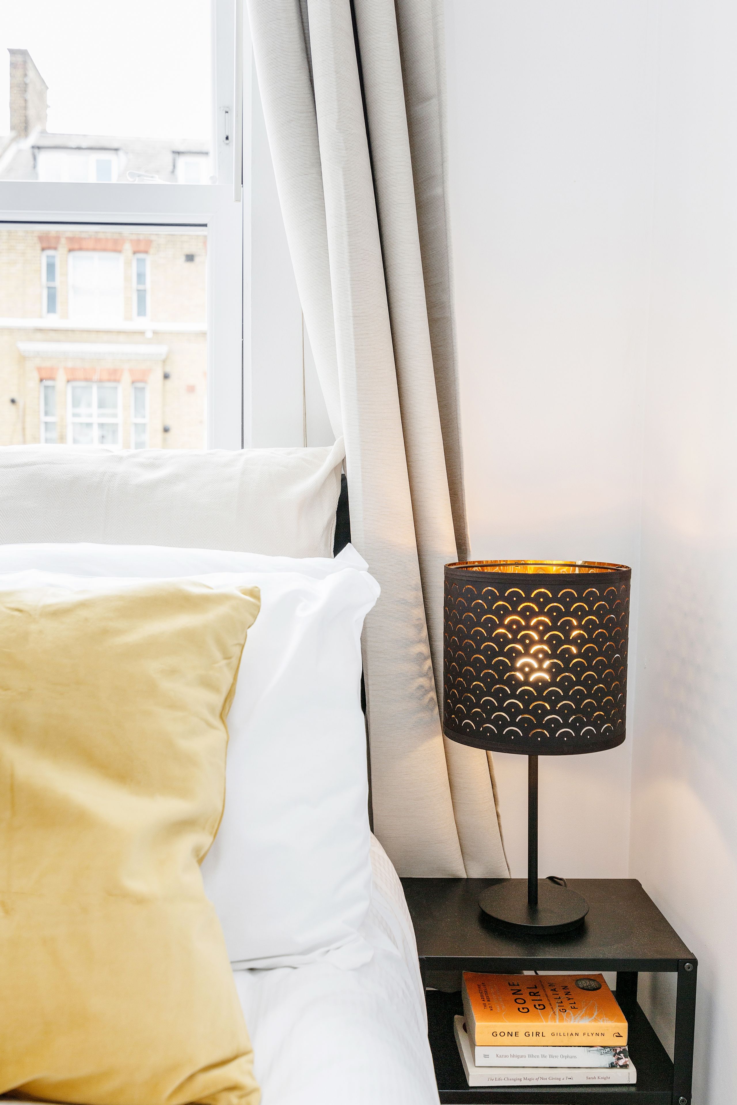 Uma cama bem arrumada com um travesseiro amarelo, uma mesa de cabeceira com uma luminária e um livro intitulado 