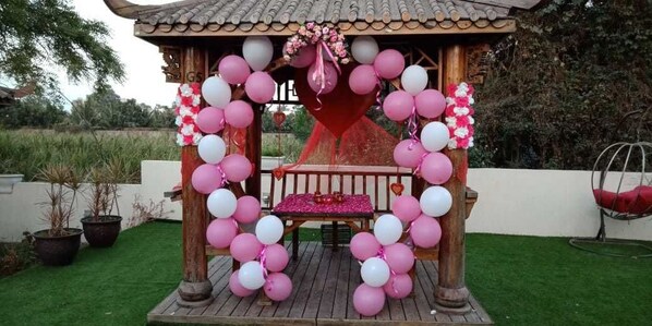 Un pabellón con un arco de globos en forma de corazón, un banco y una pequeña mesa con mantel rosa.