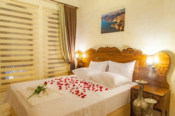 A hotel room with a wooden headboard, a bed with white linens, a bedside table with a decorative vase, and a painting of a coastal landscape on the wall.
