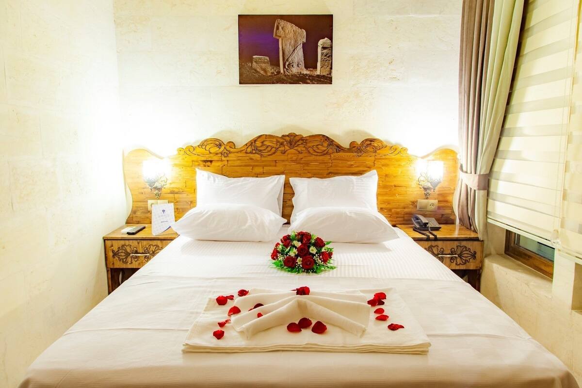 A neatly made bed with white linens, a bouquet of red roses, and rose petals arranged on a folded towel. There are two bedside tables with lamps, a wooden headboard, and a framed picture on the wall.