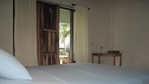 A bedroom with a bed, a wooden door, a small table with a bottle, and a view of a palm tree outside.