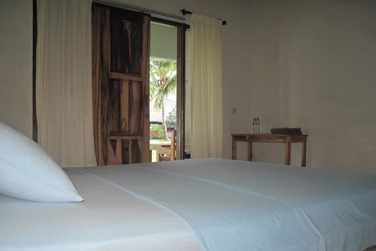 A bedroom with a bed, a wooden door, a small table with a bottle, and a view of a palm tree outside.