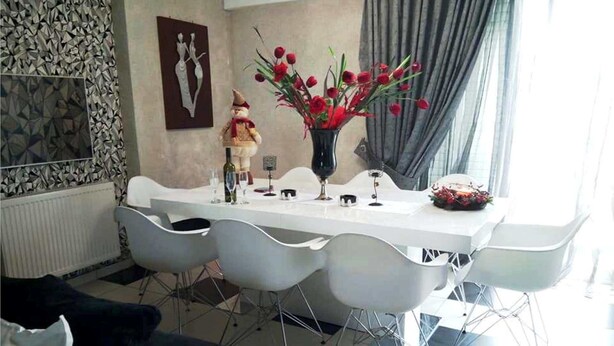 En spiseplads med et hvidt bord dækket til fire personer, omgivet af moderne hvide stole. En vase med røde blomster står på bordet, og der hænger et indrammet kunstværk på væggen bagved.