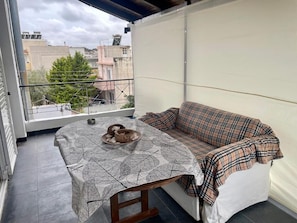 Un balcón con una mesa cubierta con un mantel estampado de hojas, un sofá a cuadros y vistas a edificios y árboles.