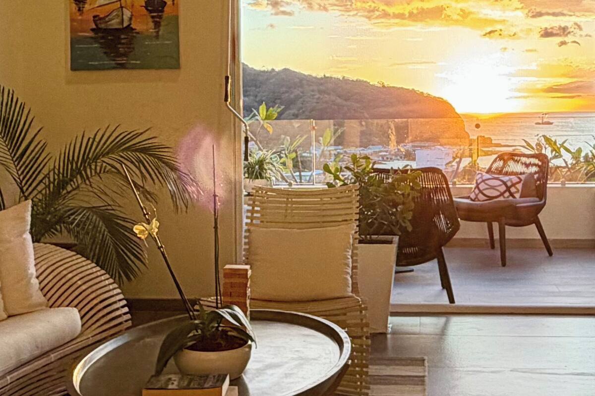 Een woonkamer met een bank, een bijzettafeltje met een plant en uitzicht op de zee bij zonsondergang.