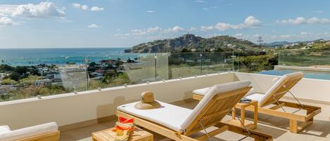 A rooftop terrace with wooden lounge chairs, a small table with fruit, and a view of the sea and distant hills.