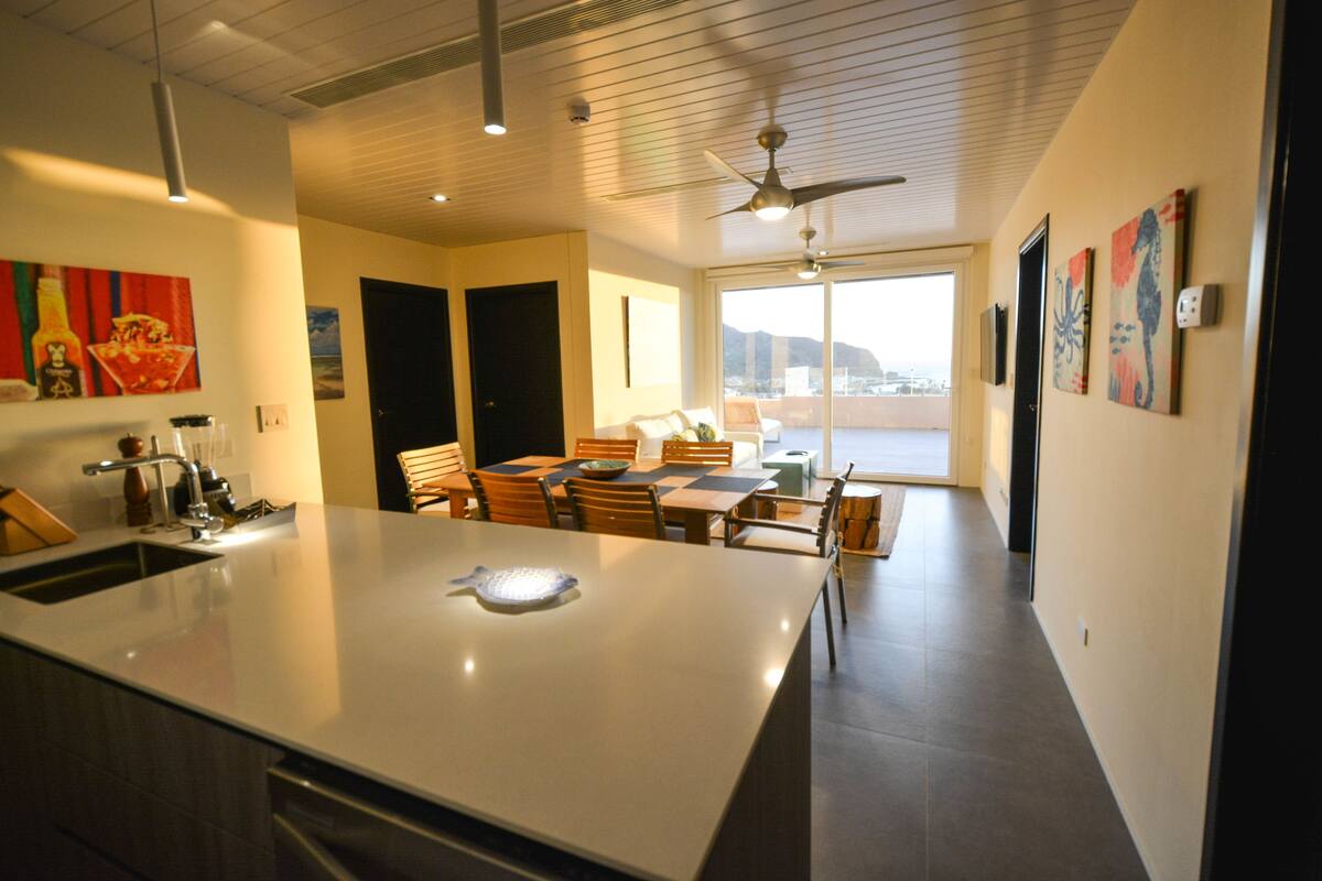 Een moderne keuken met een groot eiland, een eethoek met stoelen en een uitzicht op de stad door grote ramen.