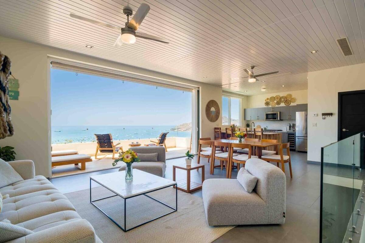 Een moderne woonkamer met een groot raam dat uitkijkt op een strand, een eethoek en een keuken.