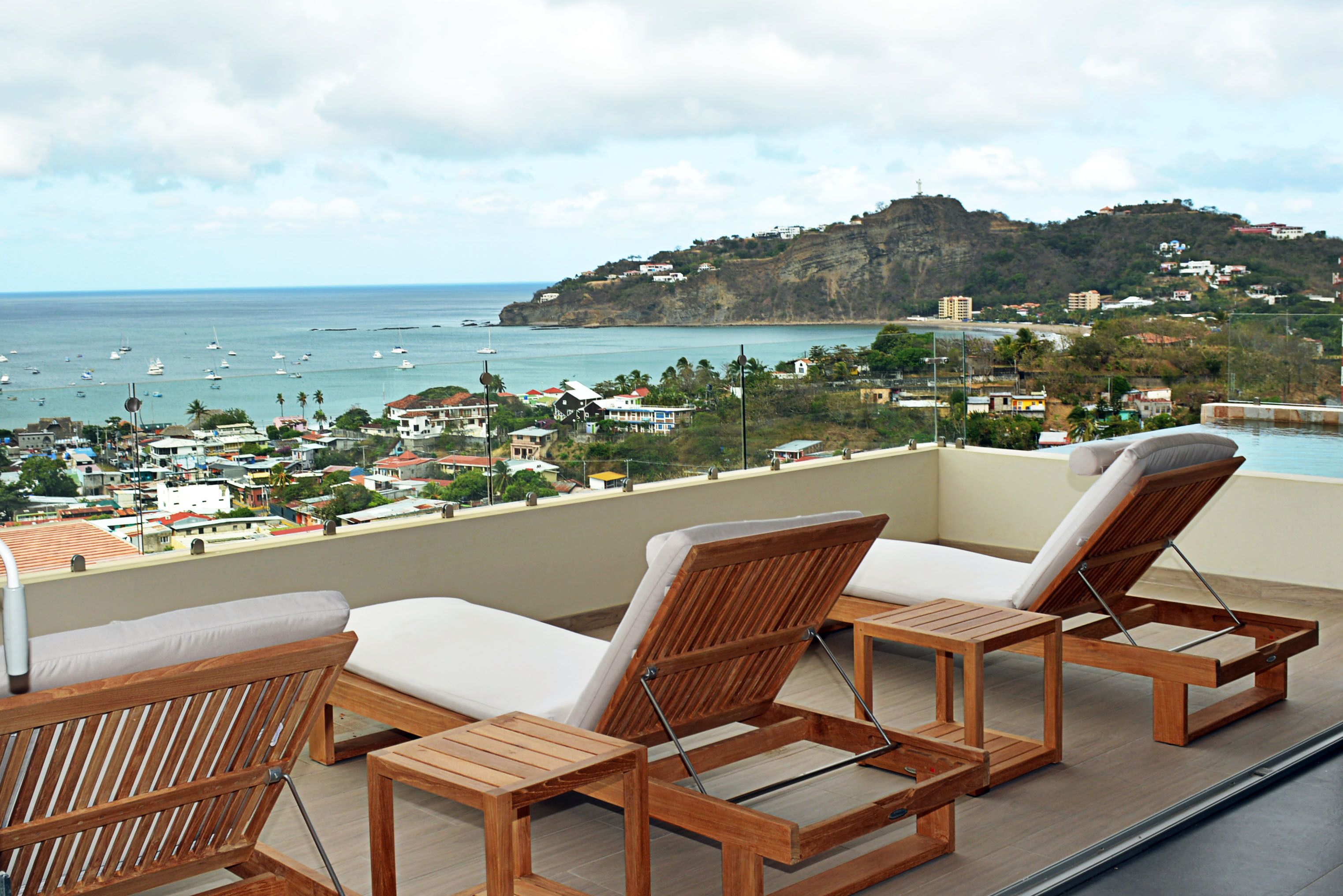 A balcony with wooden lounge chairs and a view of the ocean and a hillside town.