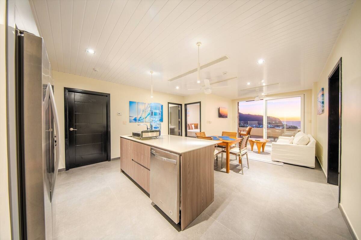 Een moderne keuken met een centraal eiland, een eethoek met een tafel en stoelen, en een zithoek met een bank en een groot raam.