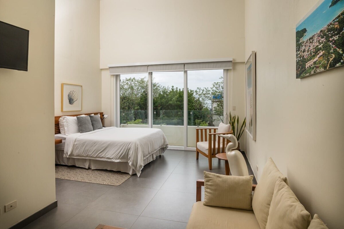 A bedroom with a bed, a chair, a sofa, a window, and a painting.