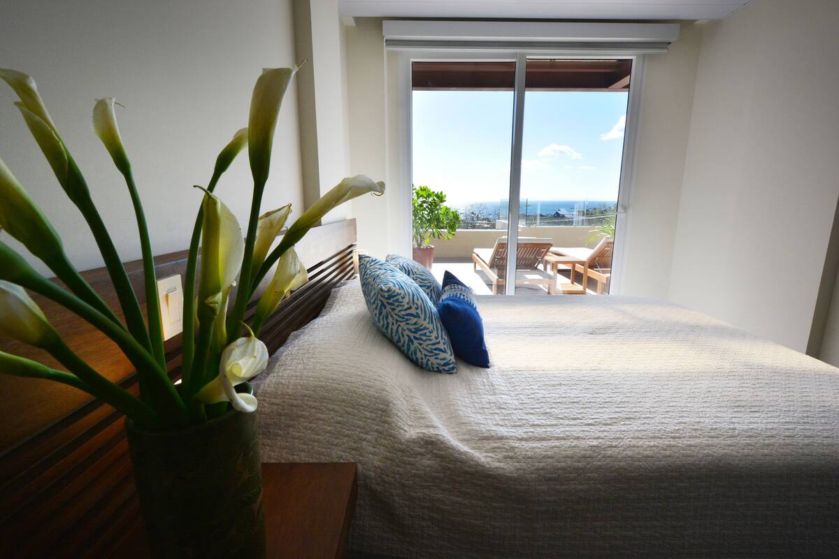 Een slaapkamer met een bed, een nachtkastje, een vaas met bloemen en een balkon met uitzicht.