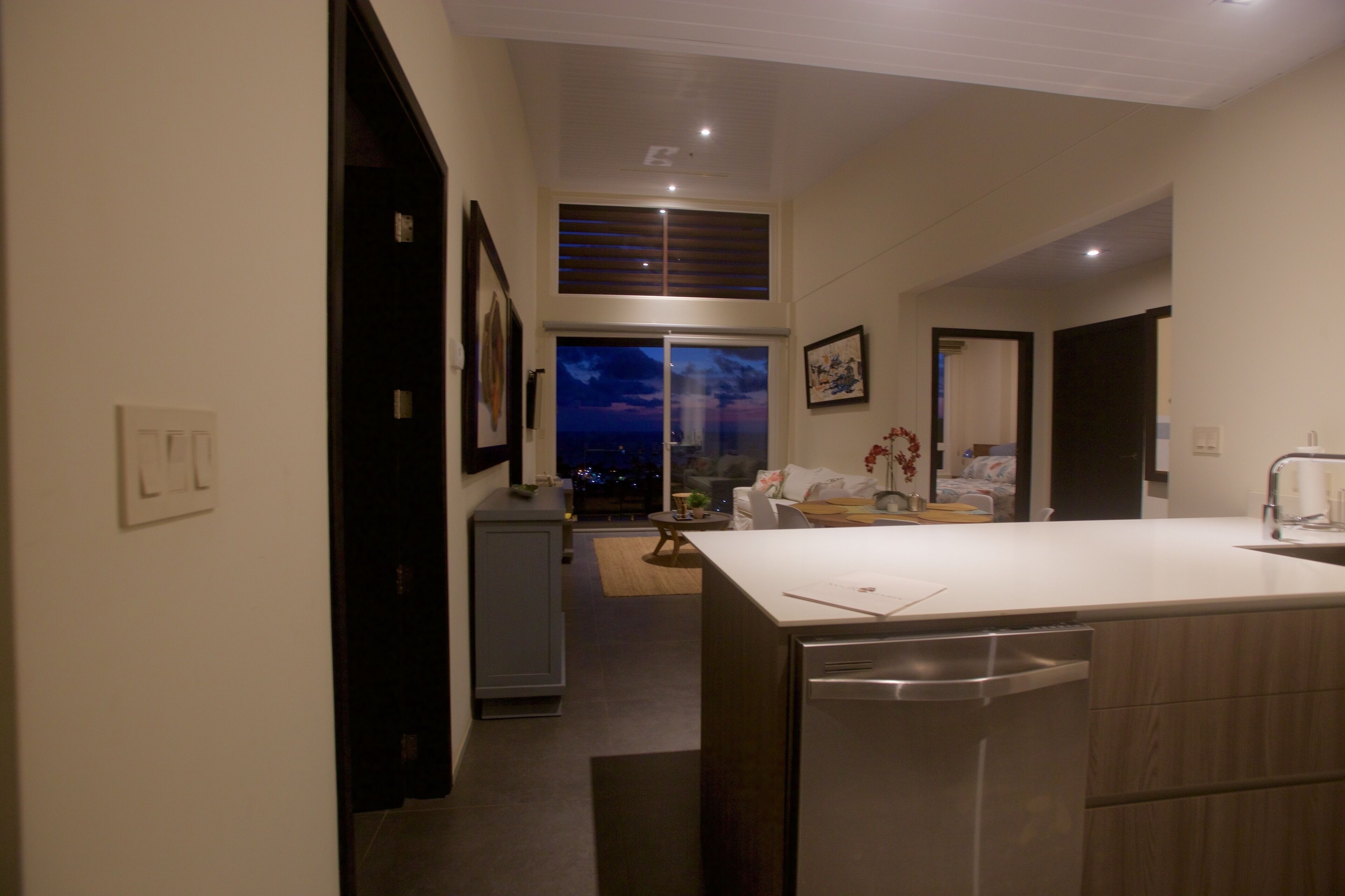 A modern kitchen with a central island, a dining area with a sofa and table, and a view of the city at night through a large window.