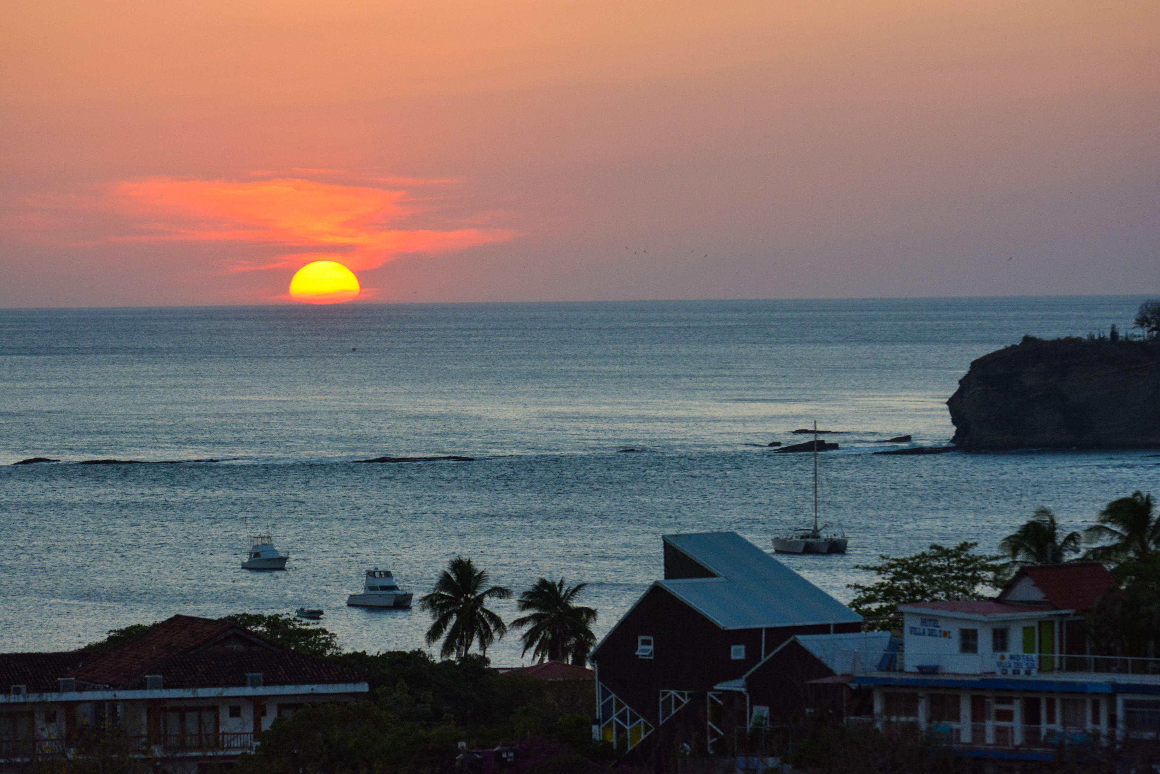 A coastal sunset view with boats, a rocky outcrop, and buildings in the foreground.
