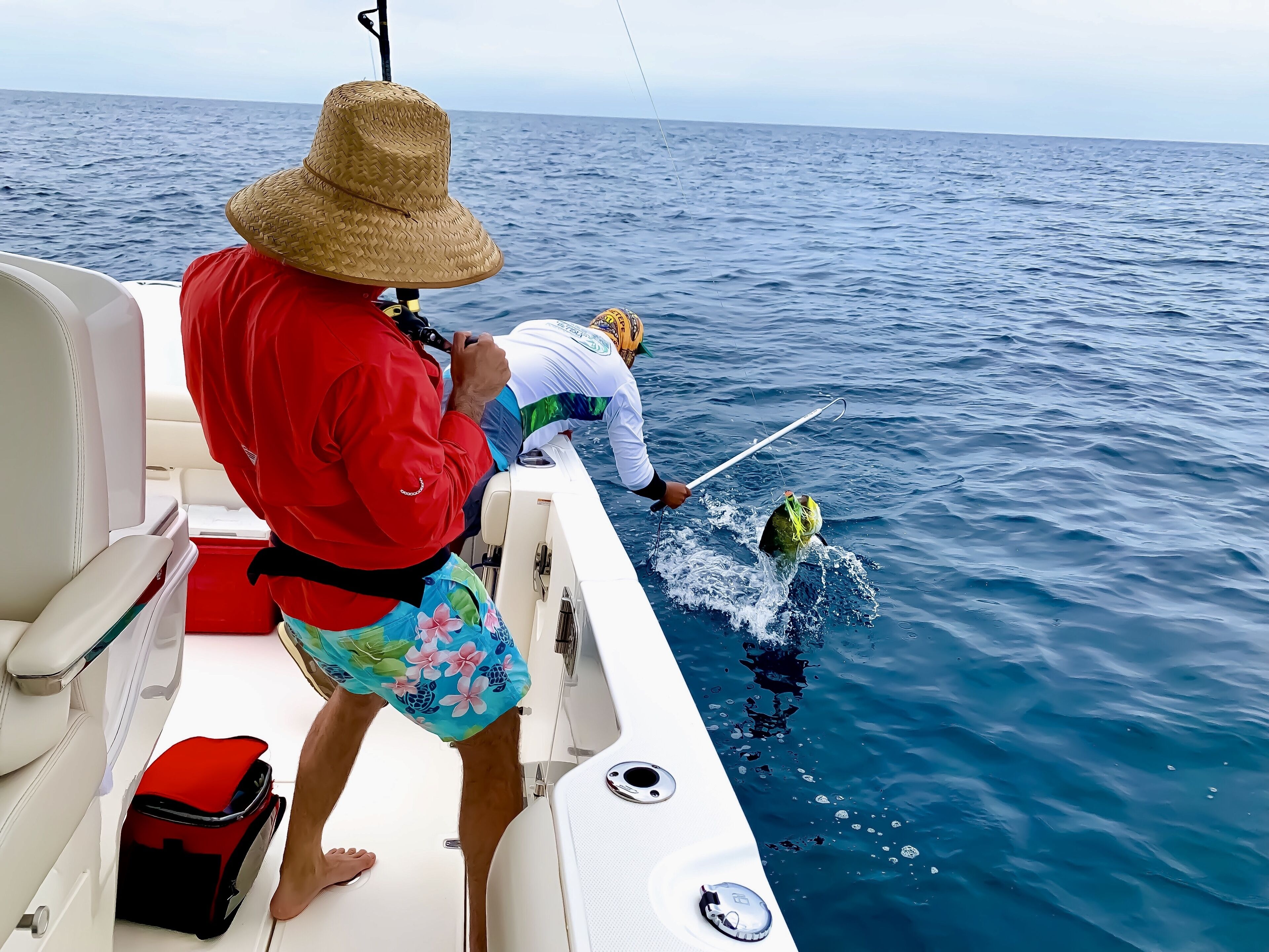 A boat with a person in a red shirt and straw hat standing at the bow, another person in a white shirt and yellow hat fishing.