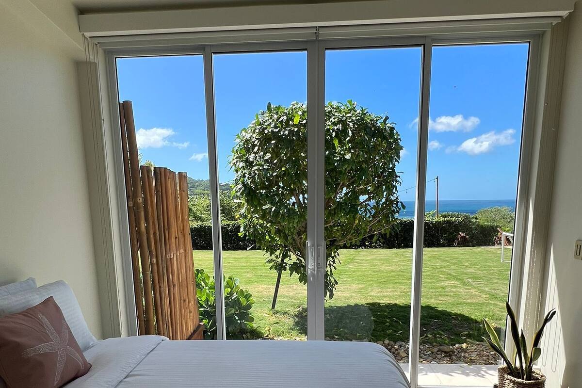 Een slaapkamer met een groot bed, uitzicht op een grasveld en een boom door een glazen schuifdeur.