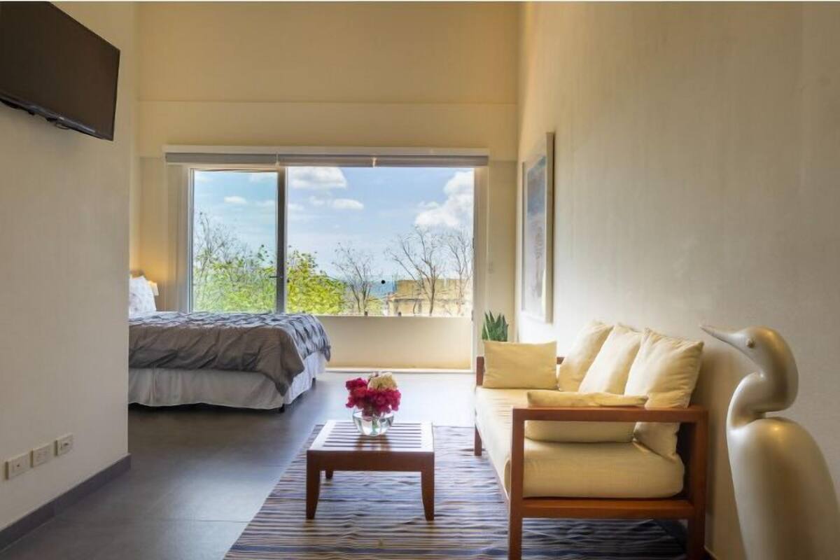 Een moderne kamer met een bed, een bank, een bijzettafeltje en een groot raam met uitzicht op bomen en gebouwen.