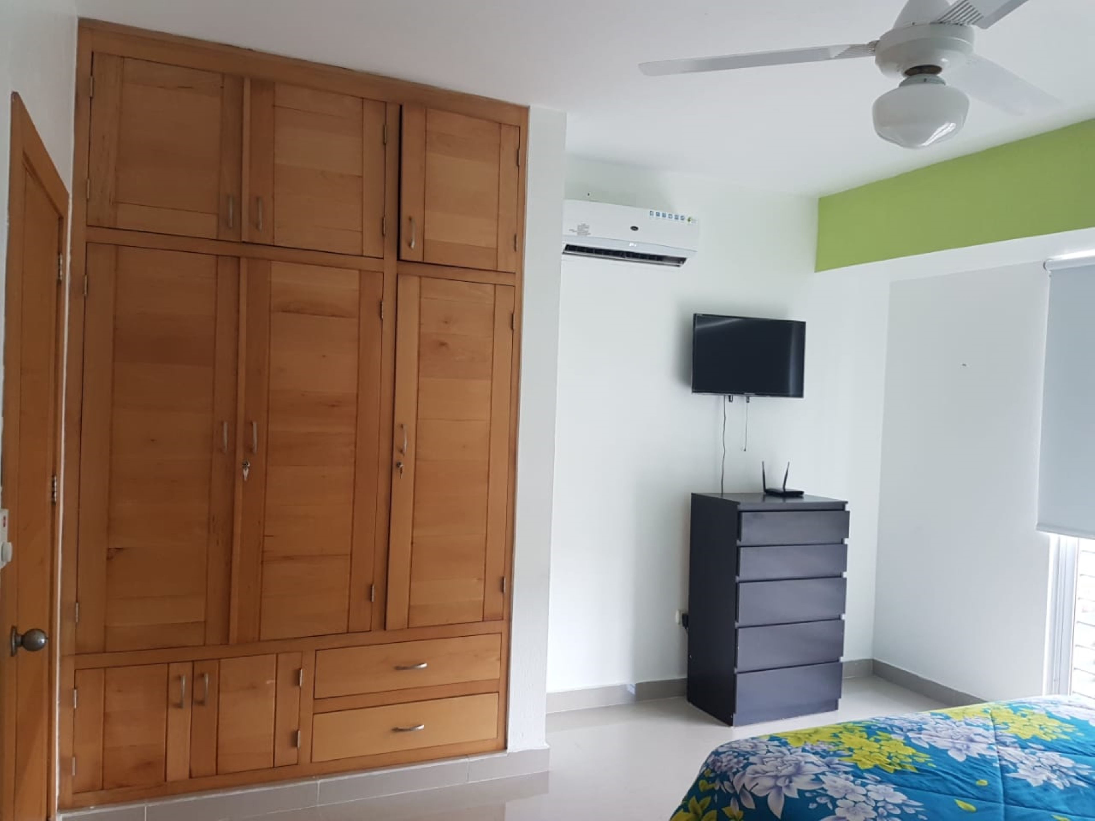 A bedroom with a wooden wardrobe, a television, a dresser, and a ceiling fan.