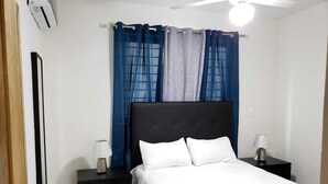 A bedroom with a bed, two bedside tables, a nightstand, a ceiling fan, and a window with blue curtains. - Amarilis Masajes (Santiago de los Caballeros)
