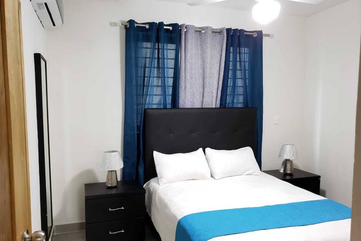 A bedroom with a bed, two bedside tables, a nightstand, a ceiling fan, and a window with blue curtains.
