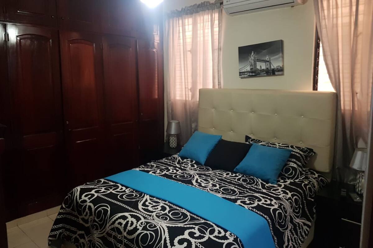 A bedroom with a bed, a ceiling fan, a wall-mounted air conditioner, and a window with curtains.