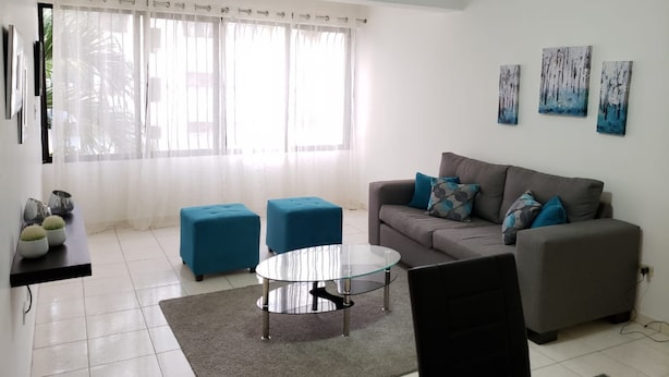 A modern living room with a grey sofa, two blue ottomans, a glass coffee table, and a ceiling fan. - Amarilis Masajes (Santiago de los Caballeros)