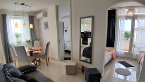 A modern hotel room with a sofa, a dining area with a table and chairs, a kitchen area, and a balcony with a view.