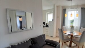 A modern living room with a grey sofa, a dining table with chairs, a kitchen in the background, and a large mirror on the wall.