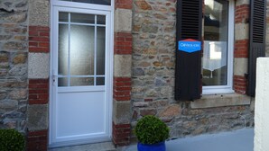 Een stenen gebouw met een witte deur en zwarte luiken, een blauw bord waarop staat dat het een vakantiewoning is, en een kleine potplant ervoor.
