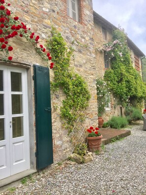 A stone building with a white door and green shutters, surrounded by gravel and potted plants.