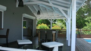 Un área de estar al aire libre cubierta con cojines blancos y muebles de mimbre, una piscina al fondo y vistas a un jardín.