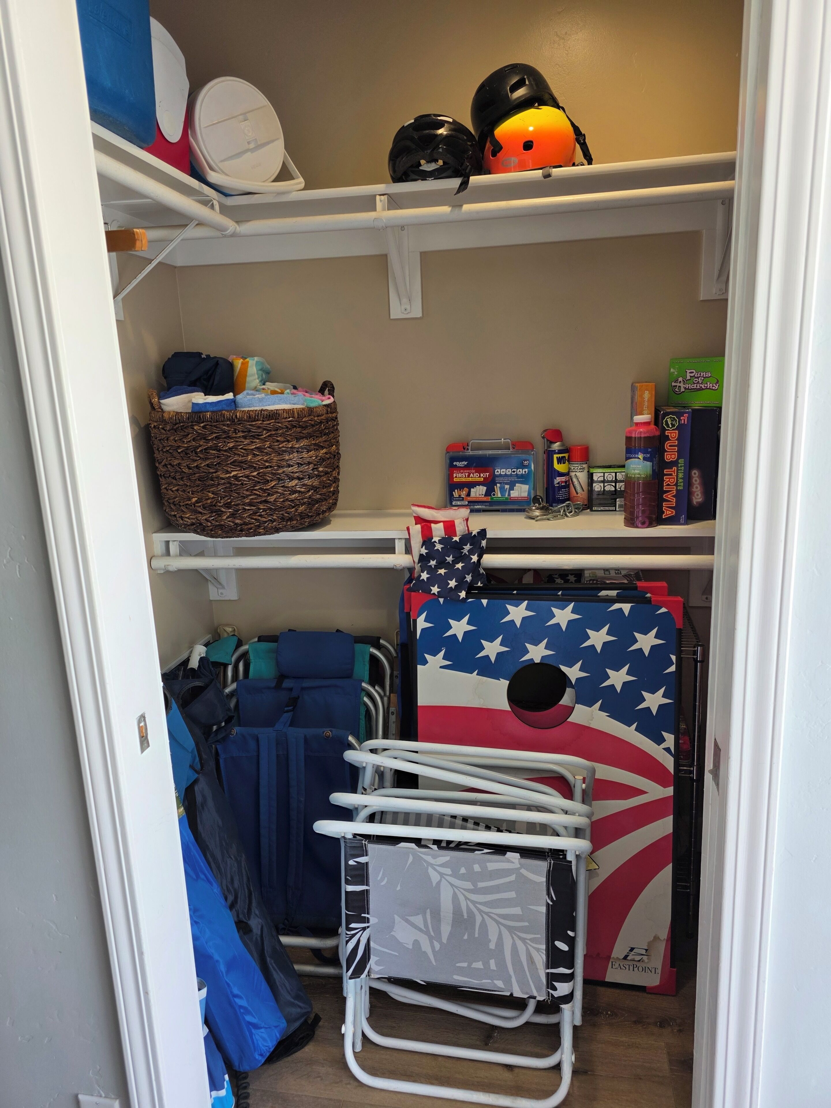 A closet with shelves containing helmets, a first aid kit, a Pub Trivia book, and a basket with clothes.
