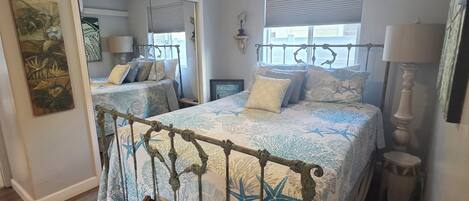 Une chambre à coucher avec un lit, un ventilateur de plafond, un miroir et une fenêtre avec des stores.
